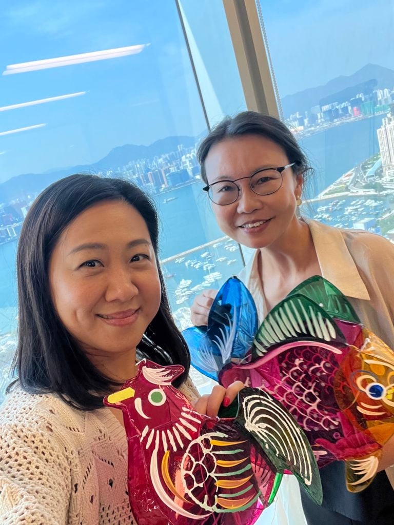 Two women smile while holding colorful rooster kites in front of a large window. The cityscape outside features tall buildings and a harbor with mountains in the background. The women wear glasses and casual clothing, standing in a bright room with natural light.
