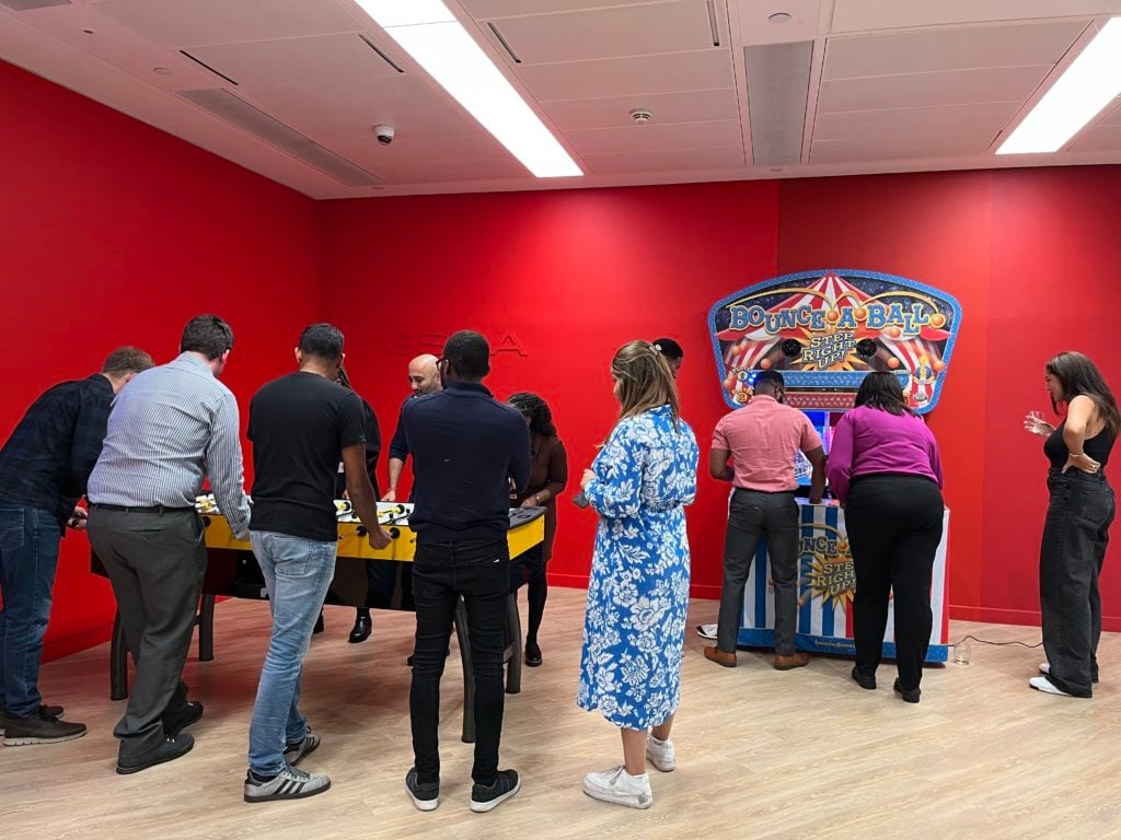 People gathered in a room with red walls, playing foosball and an arcade game. The group includes men and women, some wearing casual clothes like jeans, T-shirts, and a blue floral dress. The setting appears relaxed and social, with everyone focused on the games.