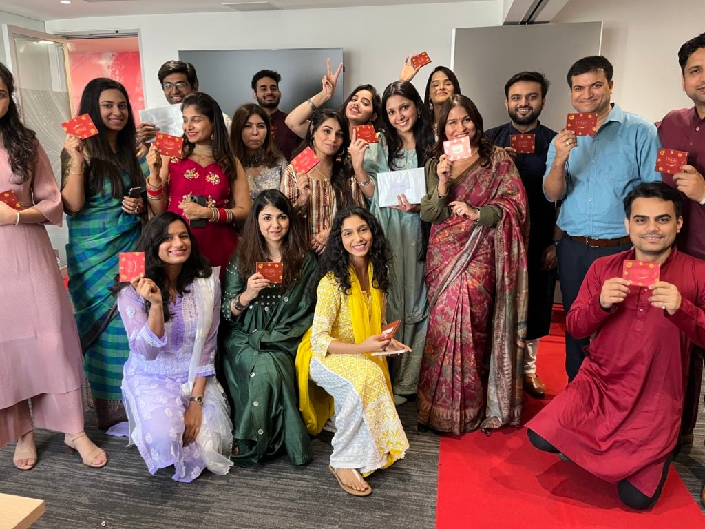 A diverse group of people in festive attire pose happily in an office setting. Most are holding red envelopes, symbolizing celebration. The group includes men and women wearing colorful traditional clothing, standing and kneeling on a red carpet. A modern office background is visible.