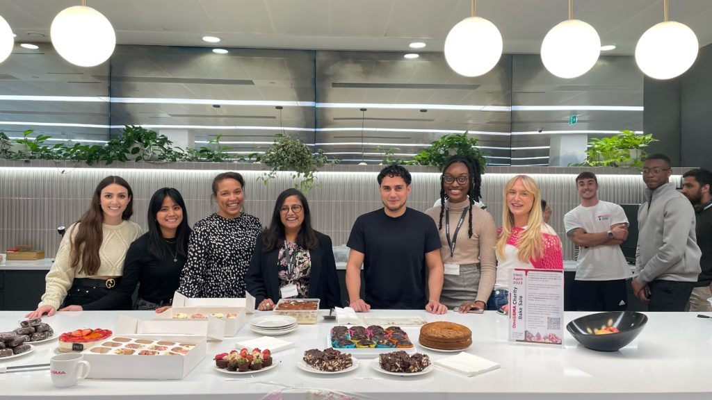 A group of eight people stands behind a table displaying a variety of baked goods, including brownies, cookies, and cakes. They smile at the camera in a modern kitchen space with greenery and pendant lights. In the background, four people engage in conversation.