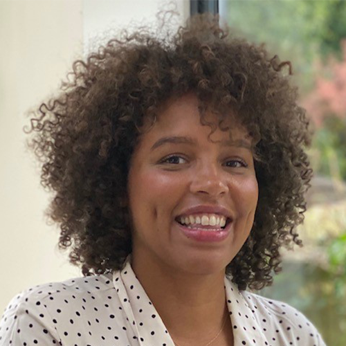 A person with curly hair is smiling warmly. They are wearing a white shirt with black polka dots. The background features a window with a blurred view of green foliage and natural light filtering in.