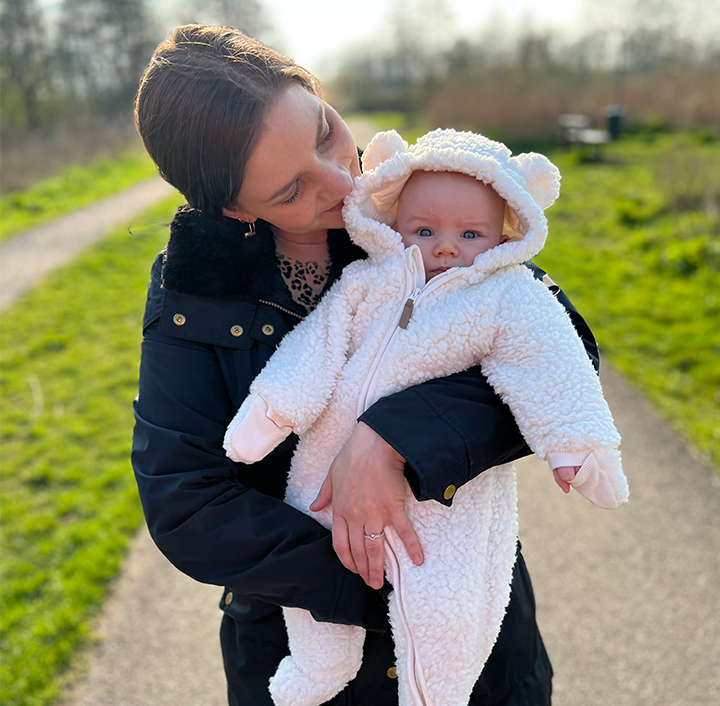 A woman with short brown hair, wearing a black jacket, gazes lovingly at an infant dressed in a fluffy, white bear-themed onesie. The baby has bright blue eyes and a hood with bear ears. They are outdoors on a sunny day, standing on a path, with grass and trees in the background.