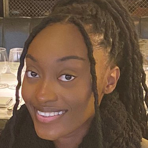 A smiling person with long, dark dreadlocks is pictured indoors. They are wearing a black sweater, with a dining setup visible in the background, including wine glasses and plates on a table. The lighting is warm, creating a cozy atmosphere.