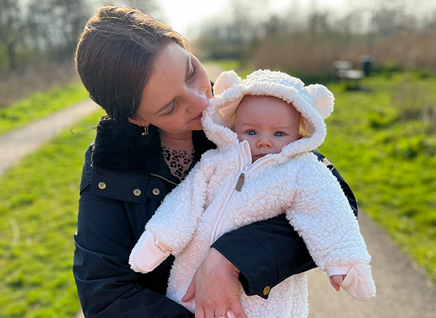 A woman holds a baby dressed in a fluffy, white coat with bear ears. They are outdoors on a path with green grass and trees in the background. The woman leans in, looking at the baby, while the baby looks at the camera with a neutral expression. It's a sunny day.