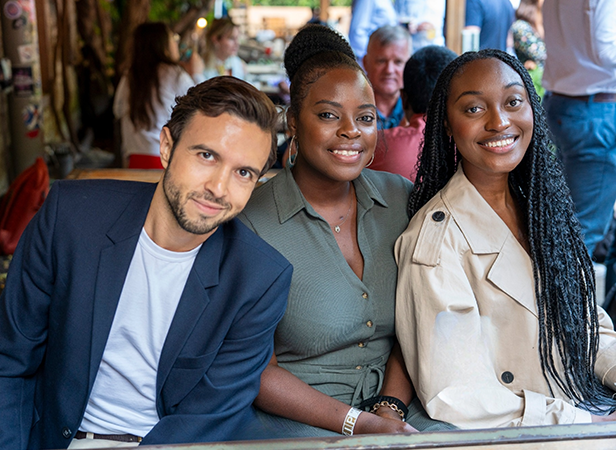 Three people are seated closely together at an outdoor gathering. The person on the left, in a dark blazer, smiles slightly. The middle individual, wearing a green shirt, smiles warmly. The person on the right in a light trench coat also smiles. Other people and greenery are in the background.