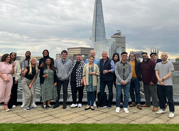 A group of sixteen people standing on a rooftop with the Shard building visible in the background. They are smiling, casually dressed, and positioned in front of a glass barrier. The sky is overcast, and there are various city buildings in the distance.