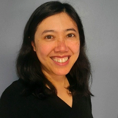 Smiling person with long dark hair wearing a black top against a gray background.