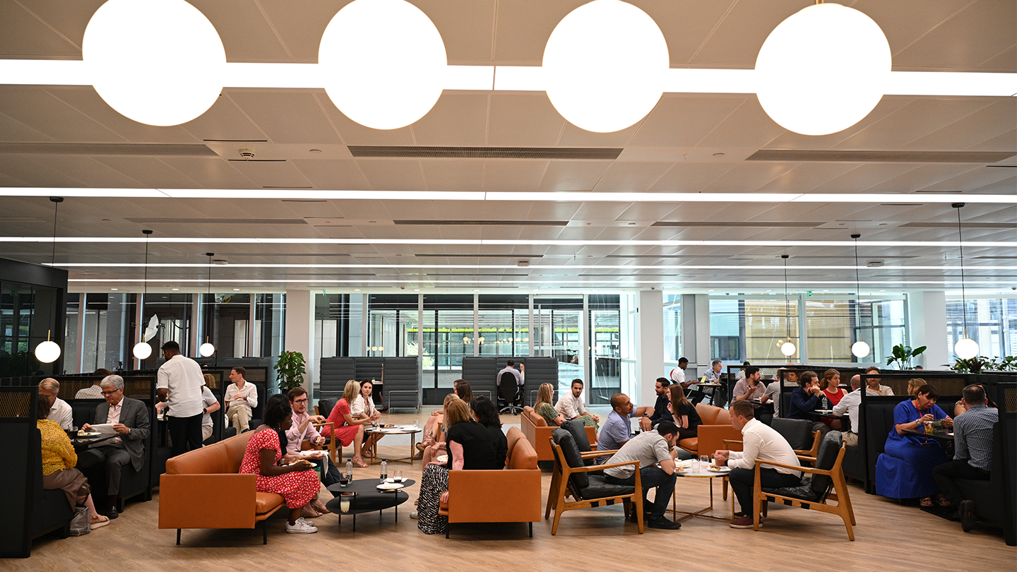 A vibrant open-plan office cafeteria where diverse people explore GSMA career opportunities while sitting and eating at small tables and lounge areas. The space features white round ceiling lights, wooden floors, and large glass windows. There are plants and a mix of orange and blue seating.