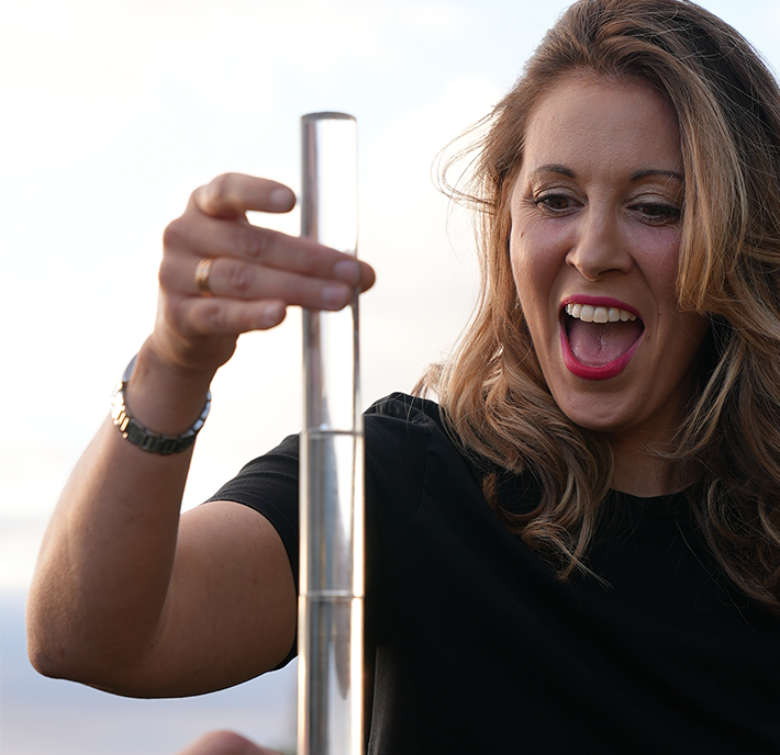 Person with long blonde hair and wearing a black shirt holds a tall, transparent measuring cylinder. She appears excited and is looking closely at the object. The background is a soft blur, likely a cloudy sky.