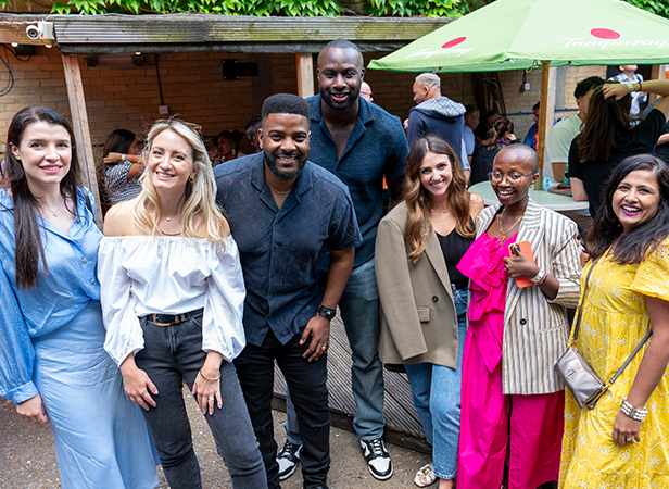 A group of seven people is outside, smiling and posing for a photo. They are dressed in a mix of casual and stylish attire, standing on a patio. In the background, there are other people, green foliage, and a large green umbrella with text on it.