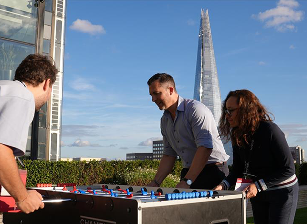 Three people play foosball outdoors on a sunny day. A man and woman are on one side, while another man is on the opposite side. The Shard skyscraper is visible in the background. Green hedges surround the area, and the sky is blue with a few clouds.