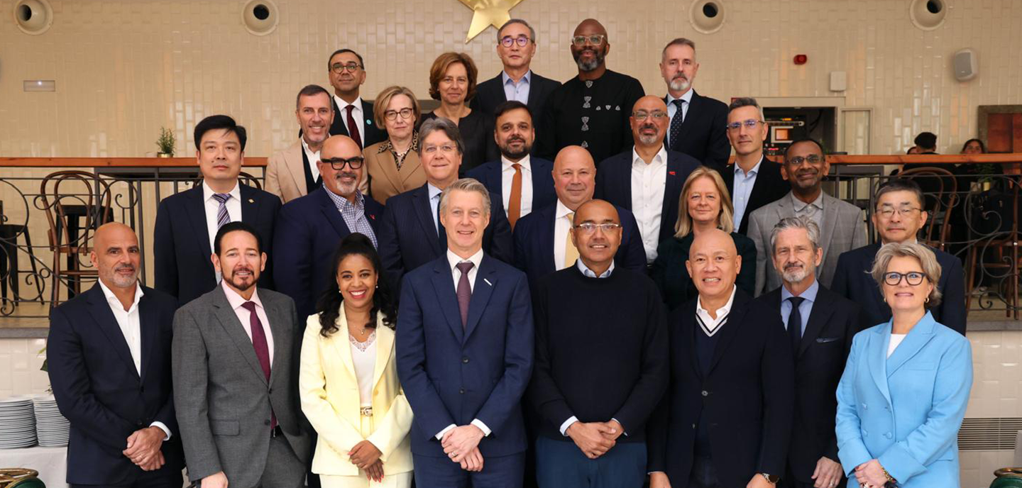 A diverse group of 23 professionally dressed people, including men and women, stand in three rows inside a warmly lit room. The background features cream tiles and decorative railings. The group is smiling, suggesting a positive and formal event.