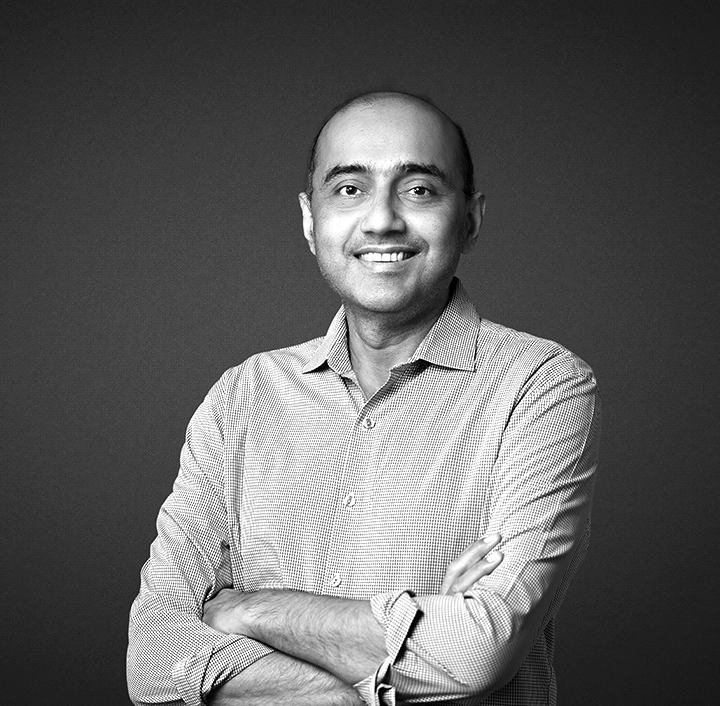 A person with short hair is smiling while standing against a dark gray background. They are wearing a long-sleeved, patterned button-up shirt with the sleeves rolled up. Their arms are crossed in front. The lighting is soft, highlighting their face. The image is in black and white.