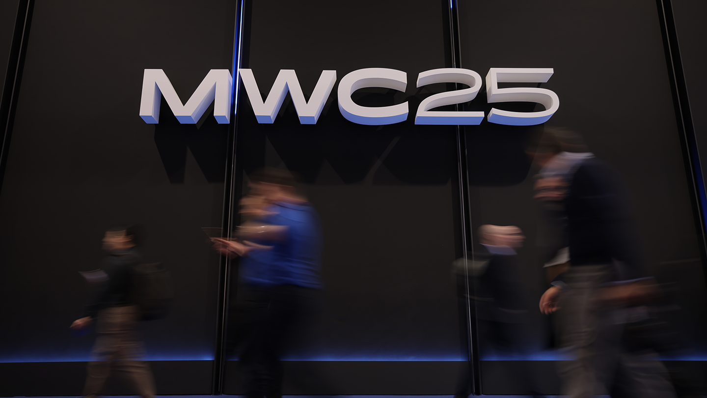 The sign reading "MWC25" glows against a dark backdrop, while blurred figures rush past. This vivid scene captures the dynamic pulse of a technology event like the Mobile World Congress, brimming with innovation and cutting-edge discussions among mobile operators.