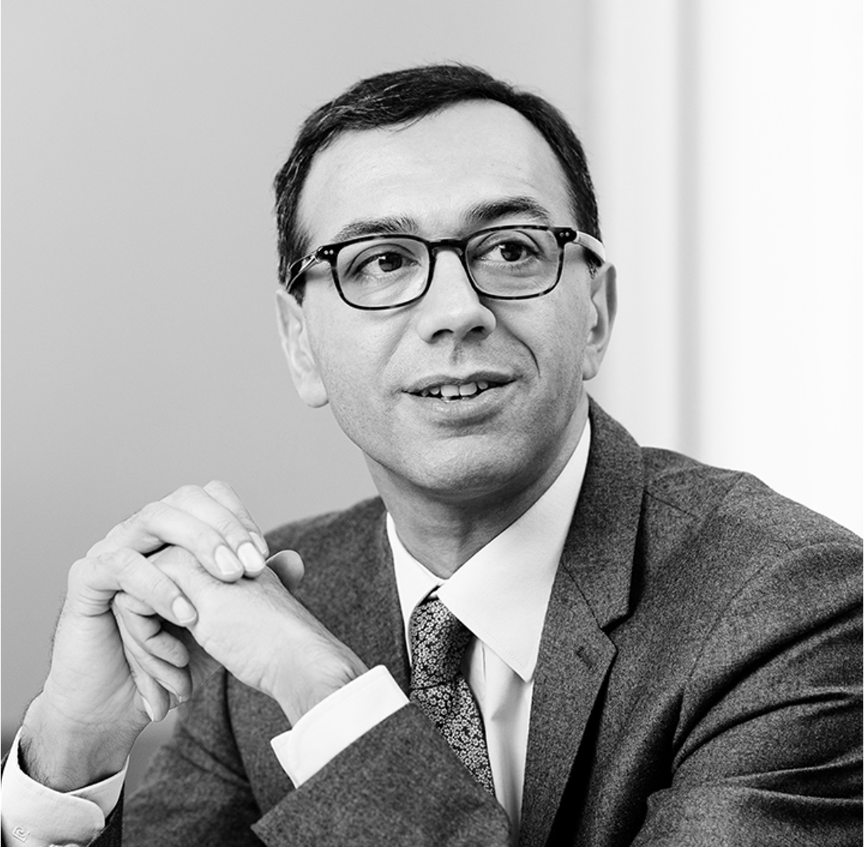 Black and white image of a person wearing glasses and a suit with a patterned tie. They are resting their clasped hands on a surface and looking slightly to the side, sitting in front of a plain background. The expression appears thoughtful, with a subtle smile.