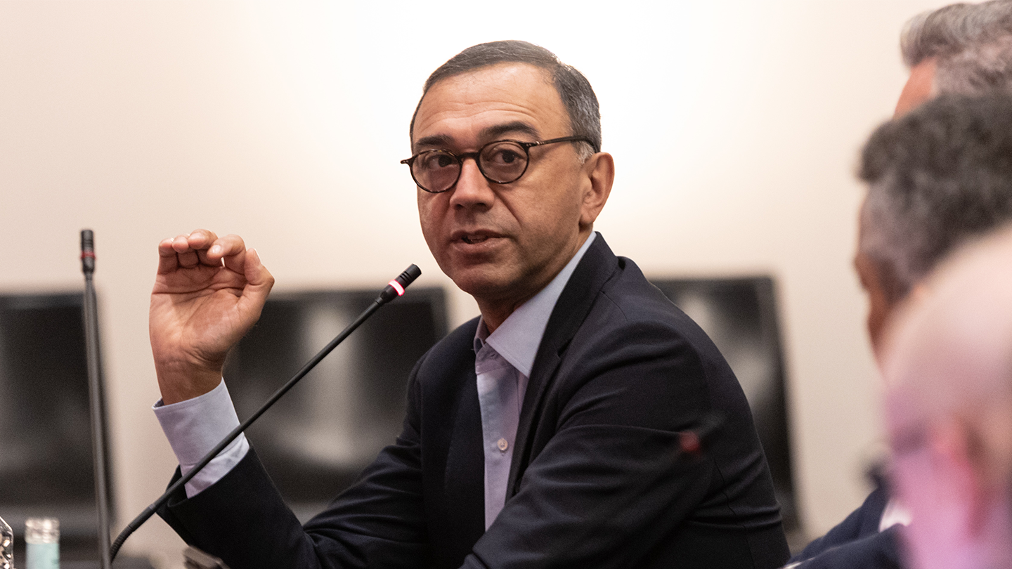 A man wearing glasses and a dark suit speaks into a microphone at a conference or meeting. He gestures with his hand while seated at a table against a blurred background.