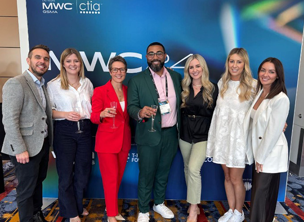 Seven people stand together smiling in front of a blue "MWC24" event backdrop. They are dressed in business or business-casual attire, with some holding drinks. The group includes men and women, all appearing happy and friendly, posing for a group photo.