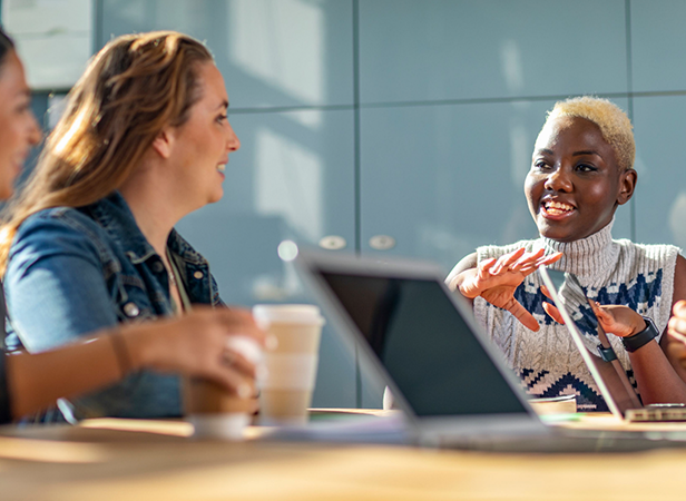 Three people sit around a table with laptops and coffee cups, engaged in a lively discussion. A smiling person with short blond hair gestures while talking, as the others listen attentively. Sunlight streams onto the table, creating a warm, collaborative atmosphere.