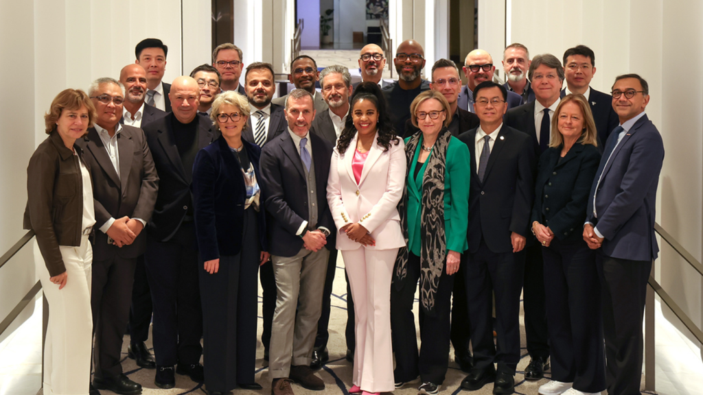 A group of 24 professionally dressed people of diverse backgrounds stand closely together indoors, smiling at the camera. The setting appears modern with white walls and a carpeted floor. One woman in the front centre wears a light pink suit.