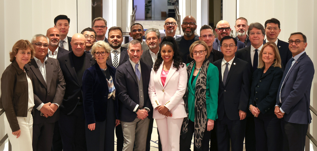 A diverse group of 24 professionally dressed people, including men and women, stand closely together indoors, smiling at the camera; the background shows white walls and a staircase.