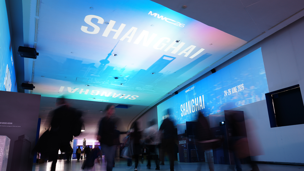 Blurred people walk in a corridor with “MWC24 SHANGHAI” and a city skyline projected onto the ceiling, capturing the dynamic energy of this year’s Mobile World Congress. Blue and purple lighting illuminates the space, while a sign on the wall displays event dates: 26-28 June 2024. Hosted by the GSMA—About the GSMA: representing more than.