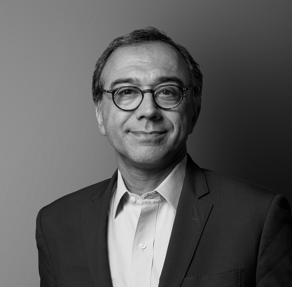 A middle-aged man with short, dark hair and glasses smiles softly. He wears a suit jacket over a buttoned-up shirt. The photo is black and white, with a plain background and soft, even lighting.