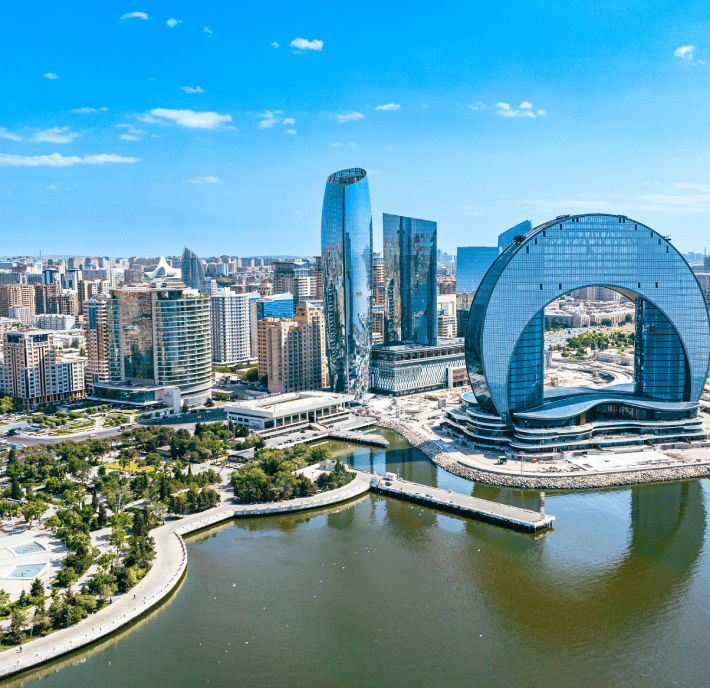 Aerial view of a modern city with unique skyscrapers by the waterfront. Notably, there is a tall, curved tower shaped like an arch. Green parks, blue skies, and scattered clouds are visible, with reflections of buildings on the calm water below.