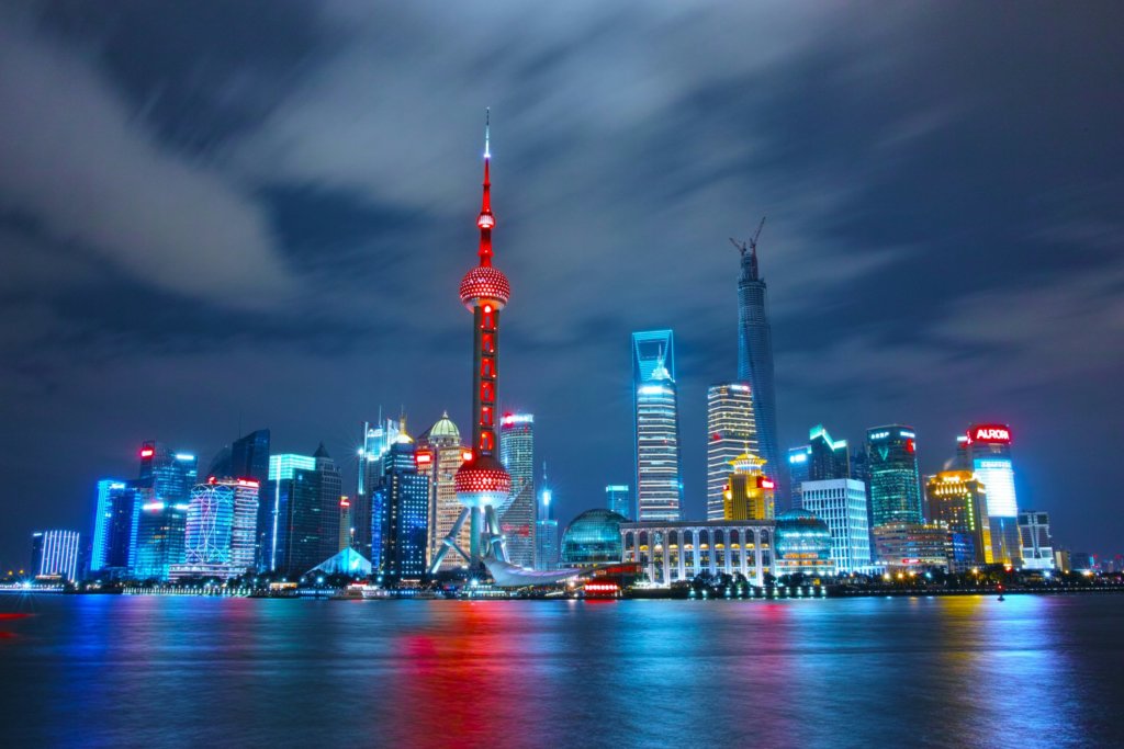 A vibrant night view of Shanghai's skyline, featuring illuminated skyscrapers reflecting on the Huangpu River. The GSMA Spectrum-enhanced Oriental Pearl Tower, with its distinctive spherical design, stands prominently lit in red. Bright lights and cloudy skies add to the lively urban atmosphere.