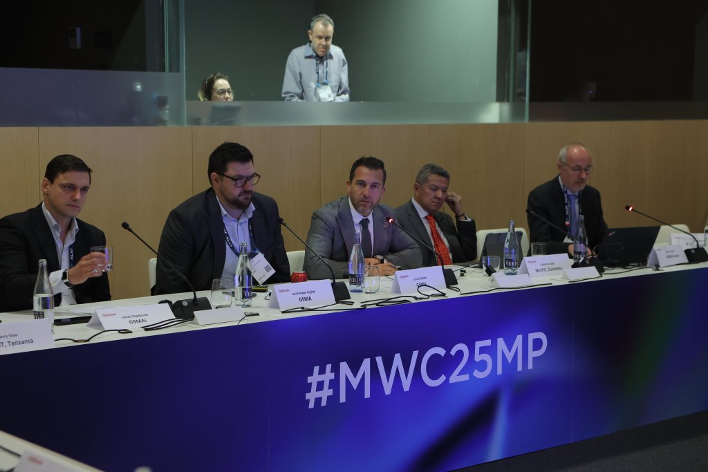Five men in suits sit at a conference table with microphones and nameplates, discussing spectrum decisions for the 5G Ministerial Programme 2025. A large banner displaying "#MWC25MP" hangs prominently. In the background, two individuals are visible behind a glass panel. Bottles of water and laptops adorn the table.