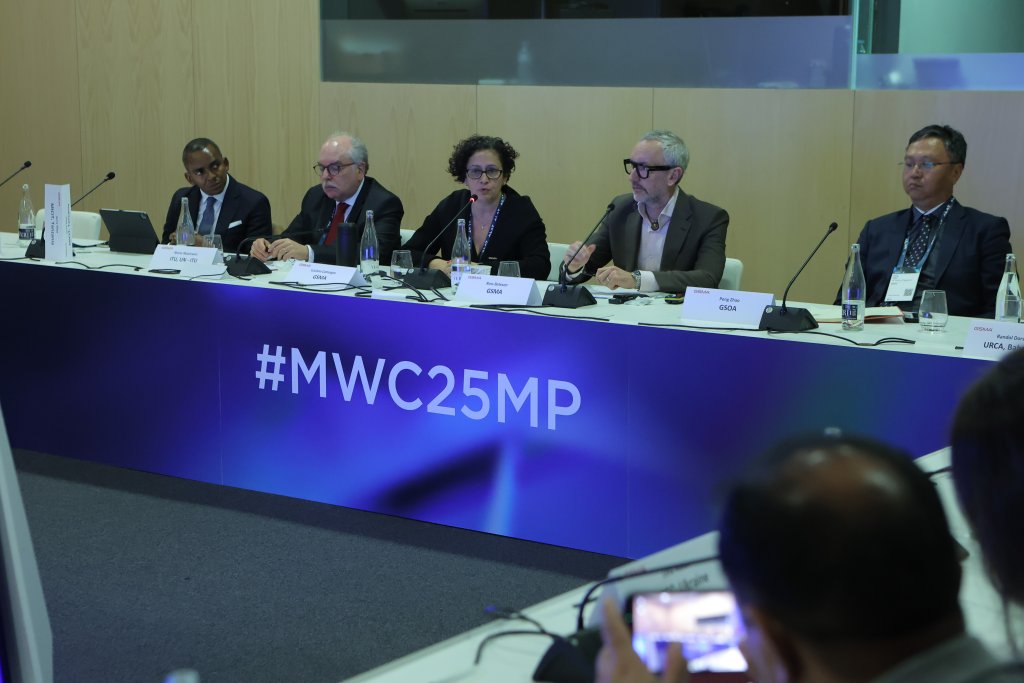 A panel of five sits at a conference table with microphones before them. A blue display reads "#MWC25MP." Wood paneling forms the backdrop. Dressed in business attire, they discuss spectrum decisions for the 5G Ministerial Programme 2025, while water bottles rest on the table.