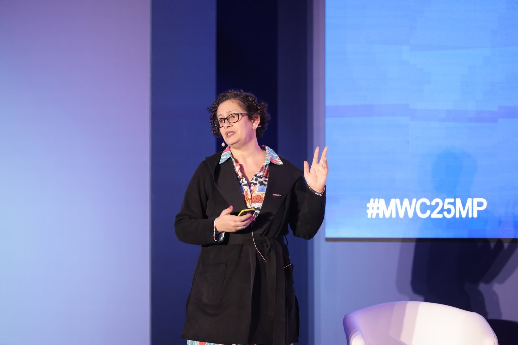 A person with short curly hair, wearing glasses and a black jacket, speaks on stage about spectrum decisions for the 5G Ministerial Programme 2025. They hold a device and gesture with their other hand against a blue backdrop displaying "#MWC25MP." A white chair is partially visible in the foreground.