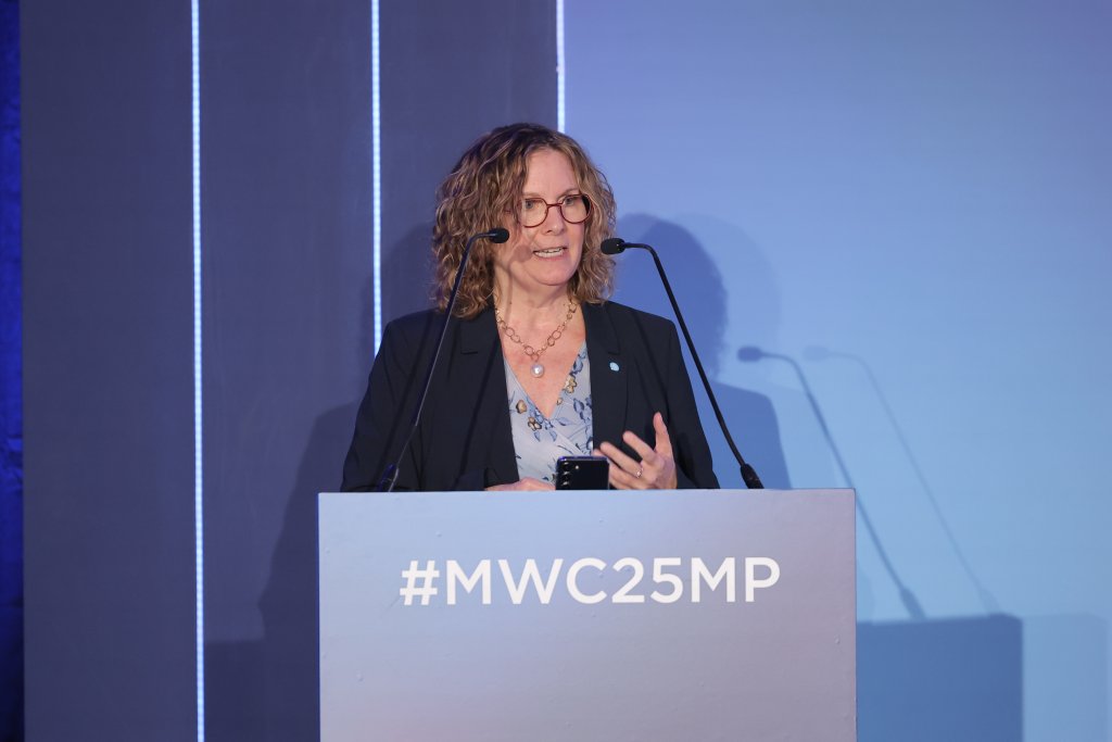 A woman with curly brown hair, wearing glasses and a navy blazer, speaks at a podium labeled "#MWC25MP," discussing spectrum decisions for the 5G Ministerial Programme 2025. Holding a phone and flanked by two microphones, she stands against a backdrop of vertical light lines on a blue wall.
