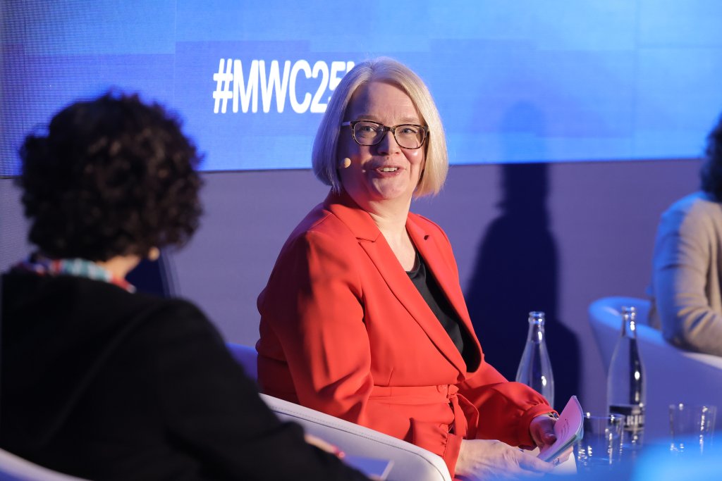 Two people are seated on stage at a discussion panel, focusing on spectrum decisions for 5G. A person with short blonde hair is wearing glasses and a red blazer, smiling while holding notes. The backdrop displays "#MWC25." A second person with dark curly hair faces away, wearing a dark jacket. Bottled water is on the table.