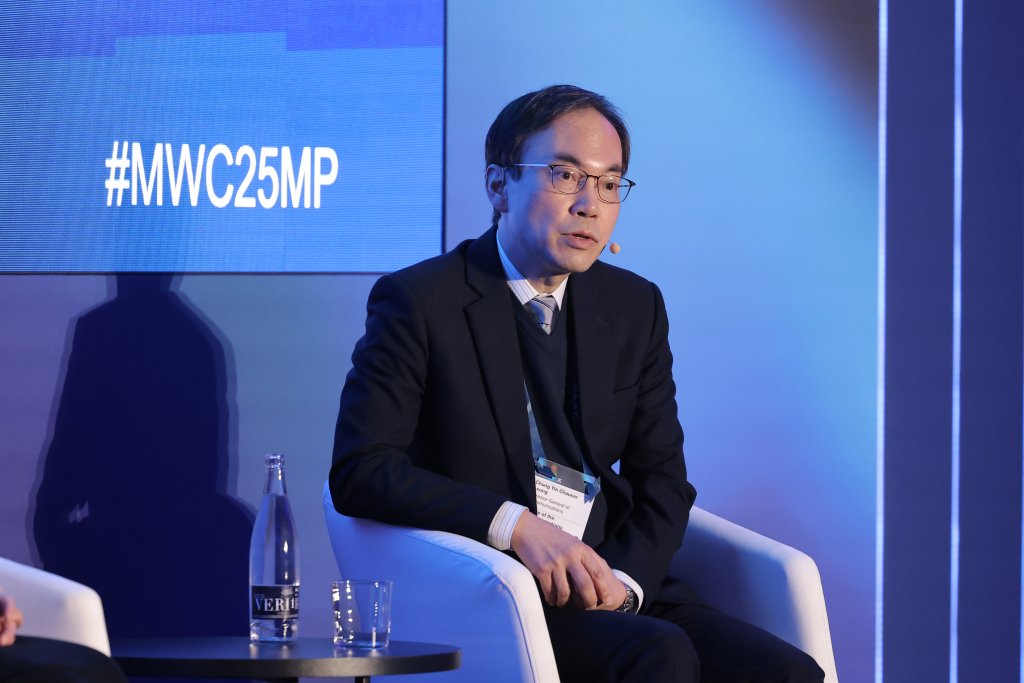 A man in a suit and glasses sits on a white chair on a stage with a blue background. Sporting a name badge, he speaks into the microphone on his face. A bottle and glass of water rest on the table beside him as the screen displays "#MWC25MP: Spectrum decisions for 5G Ministerial Programme 2025.