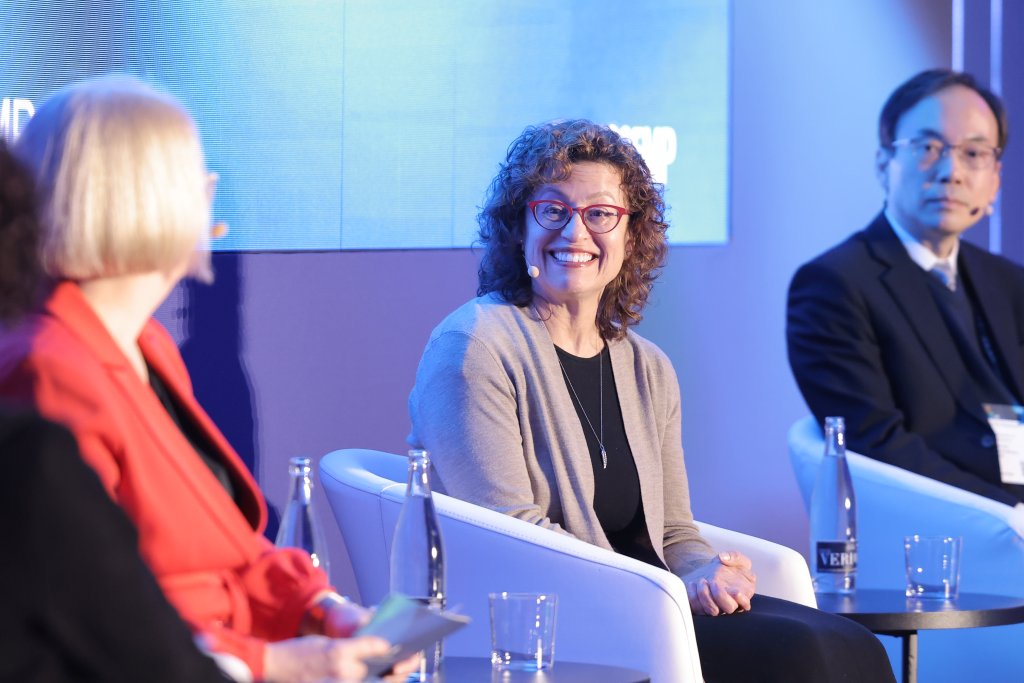 Three people are seated on stage for a panel discussion. The woman in the center, wearing glasses and a beige sweater, is smiling. The woman on the left in a red jacket and the man on the right in a suit engage as they discuss spectrum decisions for 5G at the Ministerial Programme 2025, with a large screen displaying key points behind them.