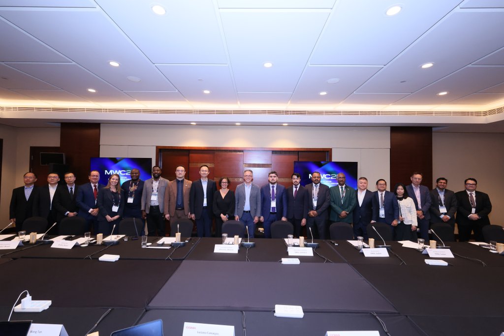 A group of 17 people in business attire stand in a conference room behind tables with microphones and nameplates. Two screens display “MWC23.” The room has bright lighting, neutral walls, and a ceiling with recessed lights.