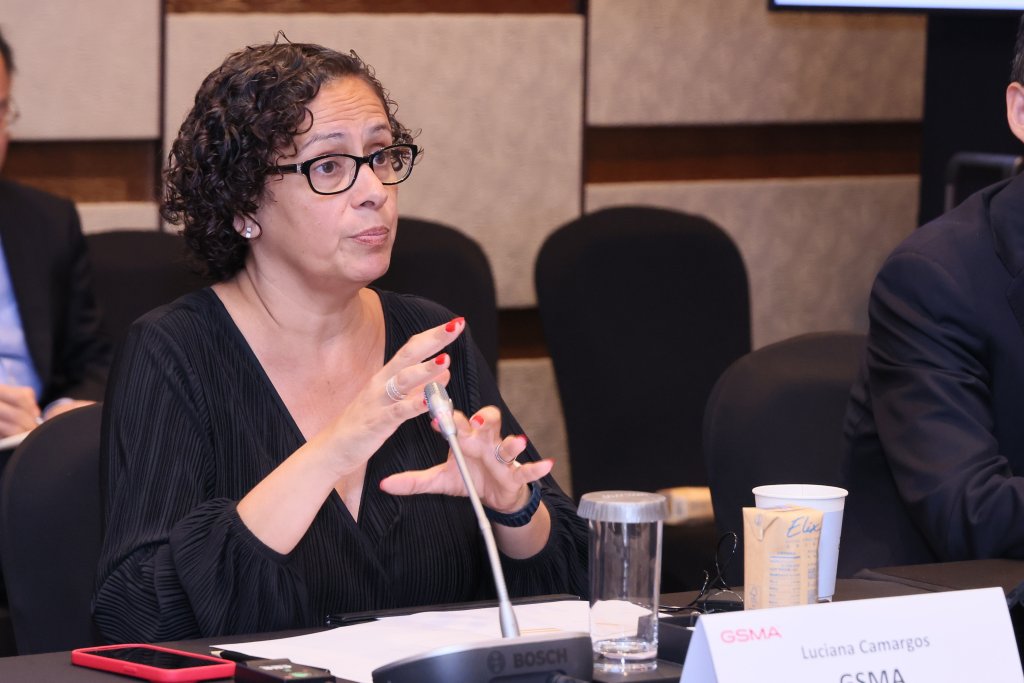 A woman with short curly hair and glasses speaks at a conference table, gesturing with her hands. She wears a black top and sits beside a microphone, a glass of water, and a paper cup. A nameplate in front of her reads "Luciana Camargos, GSMA.