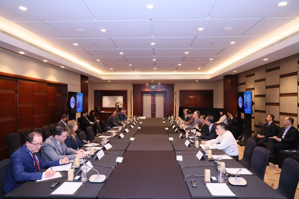 A group of people in formal attire sit around a large, U-shaped conference table in a modern meeting room. Nameplates, microphones, and notepads are on the tables. Presentation screens and an audience are visible in the background.