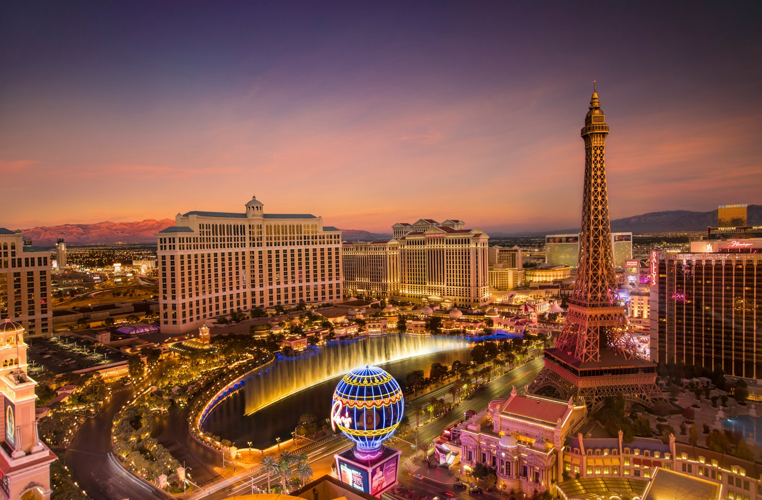 Aerial view of the Las Vegas Strip at sunset, featuring the Eiffel Tower replica at Paris Las Vegas, GSMA Spectrum lighting up casino hotels, dazzling Bellagio fountains, neon lights, vibrant skies, and distant mountains in the background.