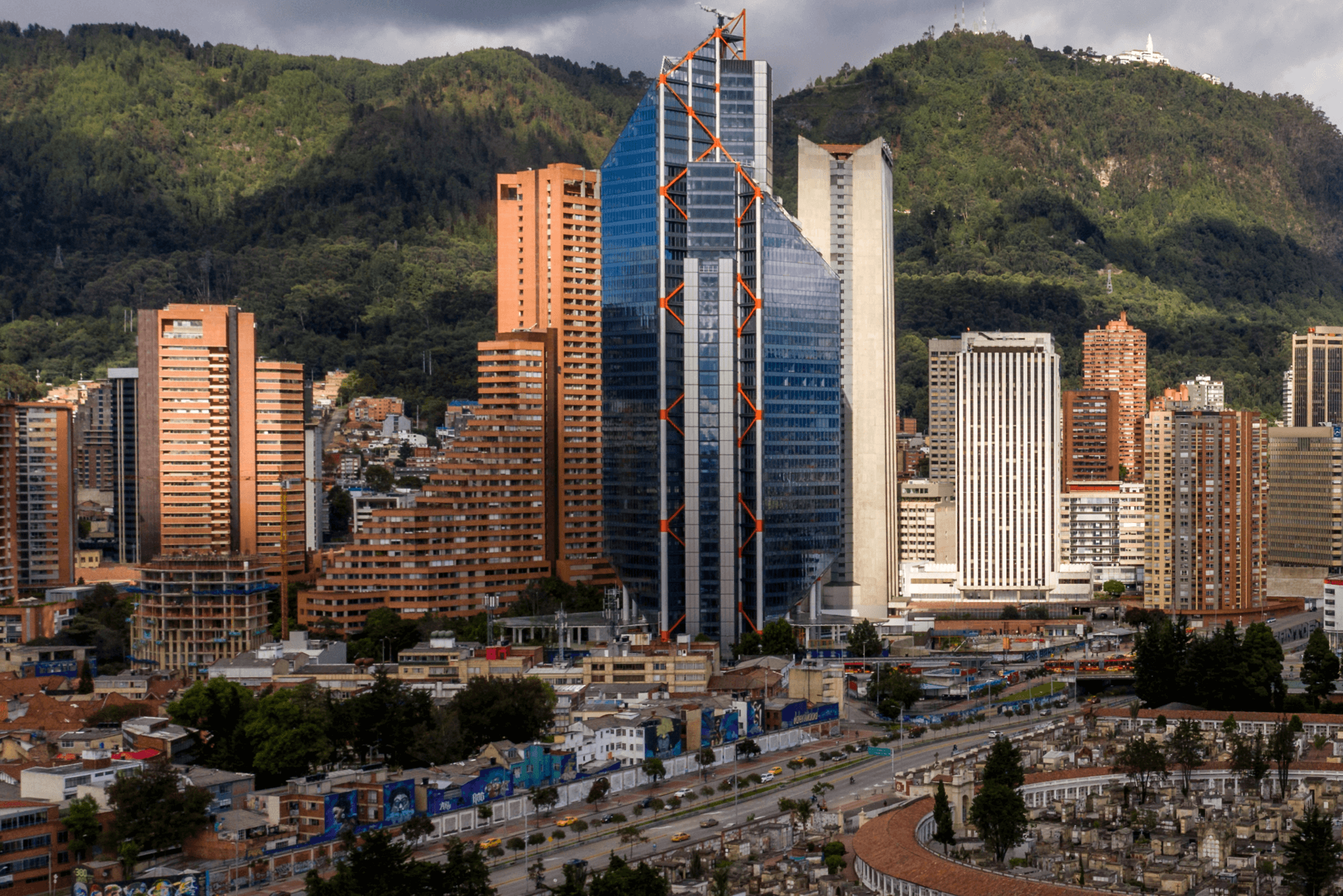 A cityscape with modern high-rise buildings set against green, forested mountains. The tallest building features a glass facade with a unique angular design and orange structural elements. In the foreground are lower buildings, a busy road, and a cemetery.