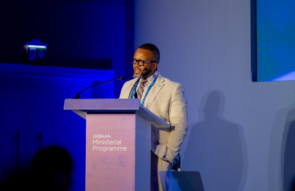 A man in a light suit and glasses speaks at a lectern labelled “GSMA Ministerial Programme” against a blue-lit background, with a microphone and lanyard visible. The scene appears to be at a formal conference or event.