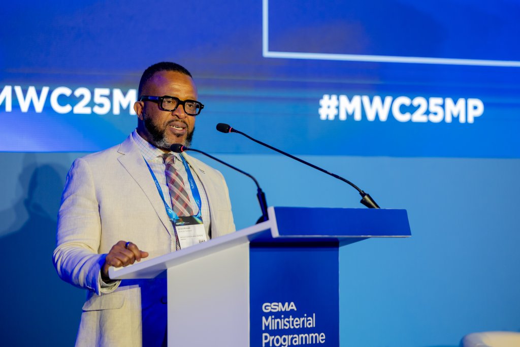 A man in a light suit and striped tie stands at a lectern, speaking into a microphone at the "GSMA Ministerial Programme," sharing spectrum takeaways from MWC Kigali. Behind him, a blue screen displays "#MWC25MP" in this formal, professional setting.