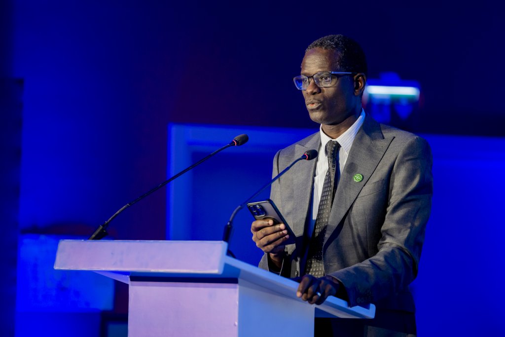 A man in a grey suit and glasses stands at a white lectern with two microphones, holding a mobile phone. He appears to be sharing Spectrum takeaways from MWC Kigali. The blue-lit background creates a formal, professional atmosphere.