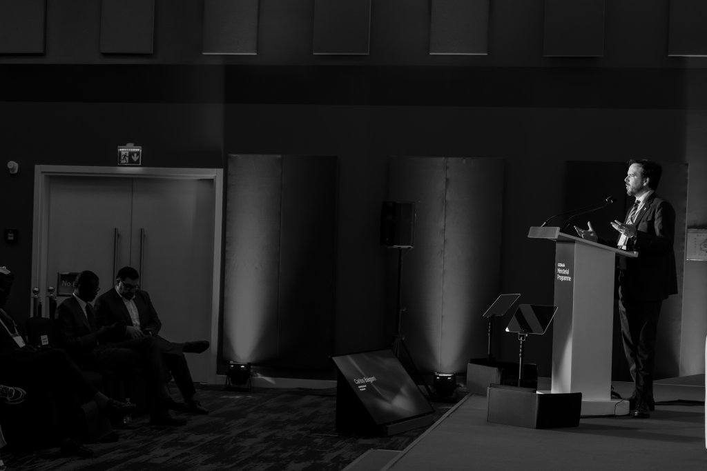A man in a suit speaks at a lectern on a stage, facing an audience seated in the dark. Spotlights illuminate the speaker, whilst the rest of the room remains dim. The background shows large panels and soft lights on the wall. The photo is in black and white.
