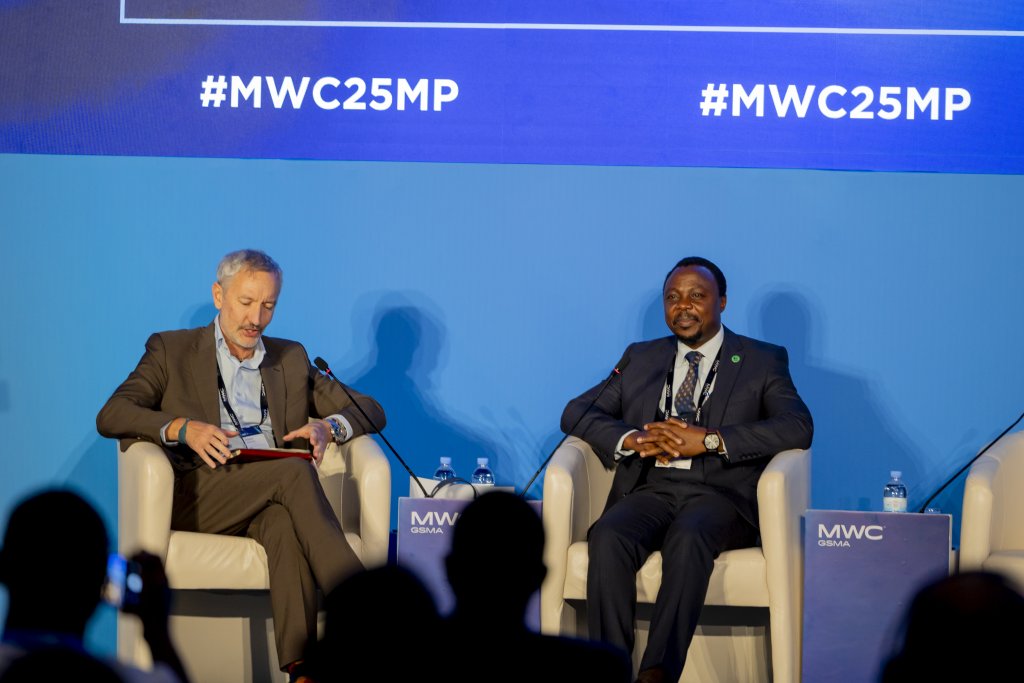 Two men in business suits sit on stage in white chairs during a panel discussion. One man holds a tablet and speaks into a microphone, whilst the other listens. A blue background displays “#MWC25MP.” Audience silhouettes are visible in the foreground.