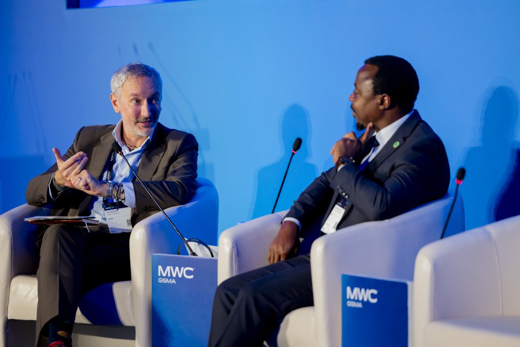 Two men in suits sit on a stage in white chairs having a conversation. They are speaking into microphones. Blue panels behind them read “MWC GSMA.” Both appear engaged, with one gesturing whilst the other listens attentively.