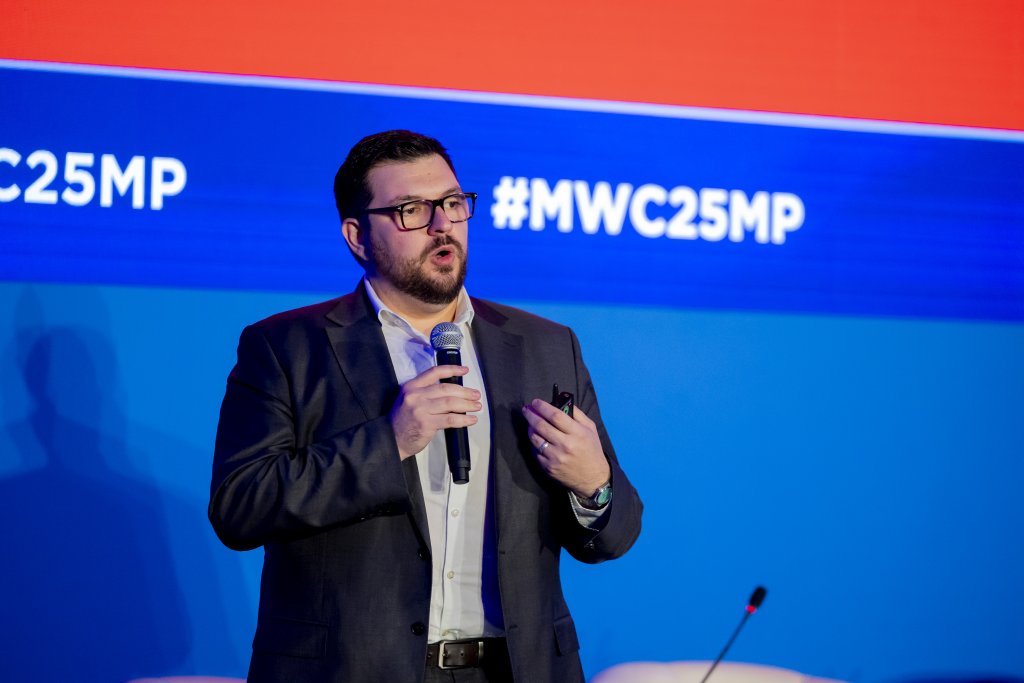 A man in a dark suit and glasses speaks into a microphone on stage. He gestures with his left hand. Behind him, a blue and red screen displays the hashtag #MWC25MP in bold white text. White chairs are partially visible at the bottom of the image.