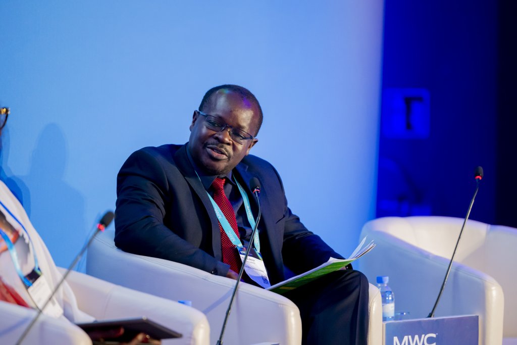 A man in a dark suit and red tie sits on a white chair, holding documents and speaking into a microphone at a conference. He faces another person off-frame. The background is blue, and a water bottle sits on a table beside him.