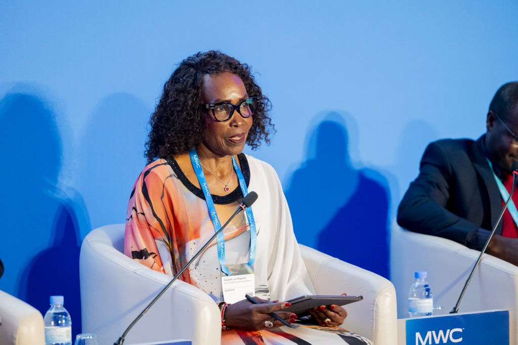 A woman with curly hair and glasses sits in a white chair, holding a tablet and speaking into a microphone. She wears a white and orange dress with a name badge. The background is blue, and water bottles and another person are visible.