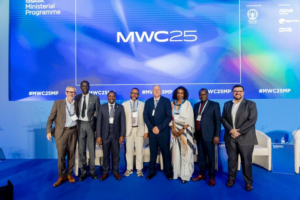 Seven people in business attire stand side by side, smiling, on a blue stage beneath a large digital display reading “MWC25.” The background includes event logos and hashtags. Two white chairs and a podium are visible on the stage.